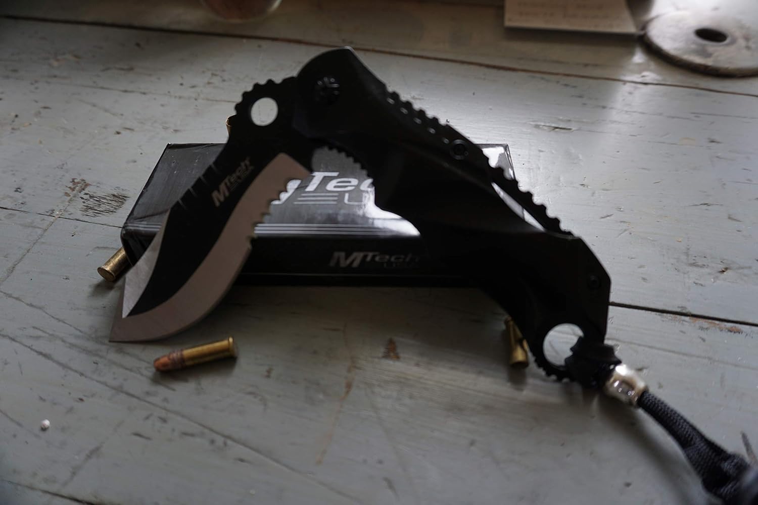 Black MTech USA MT-A808 assisted opening folding knife with a recurved partially serrated blade, jimped black handle and a paracord lanyard, resting on its box