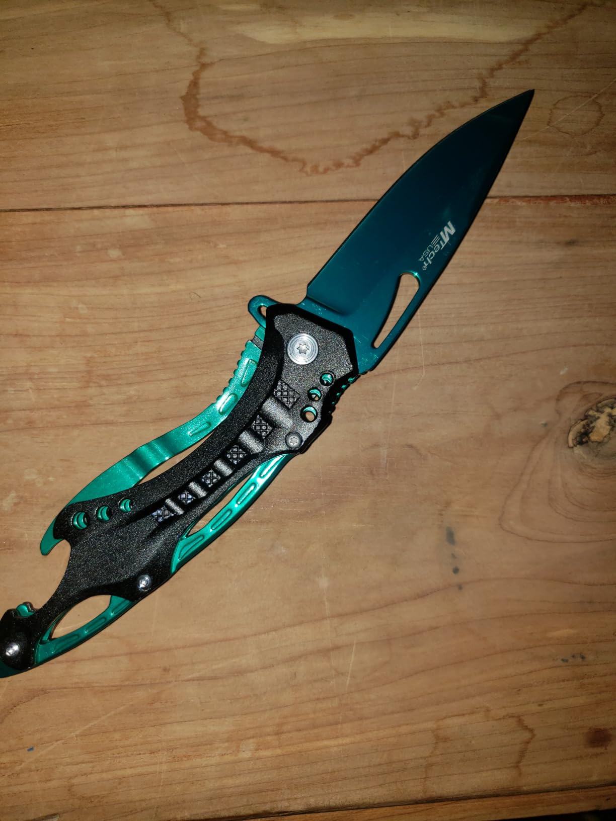Teal MTech USA Ballistic folding knife lying open on a wooden board, showing the skeletonized handle and thumb slot