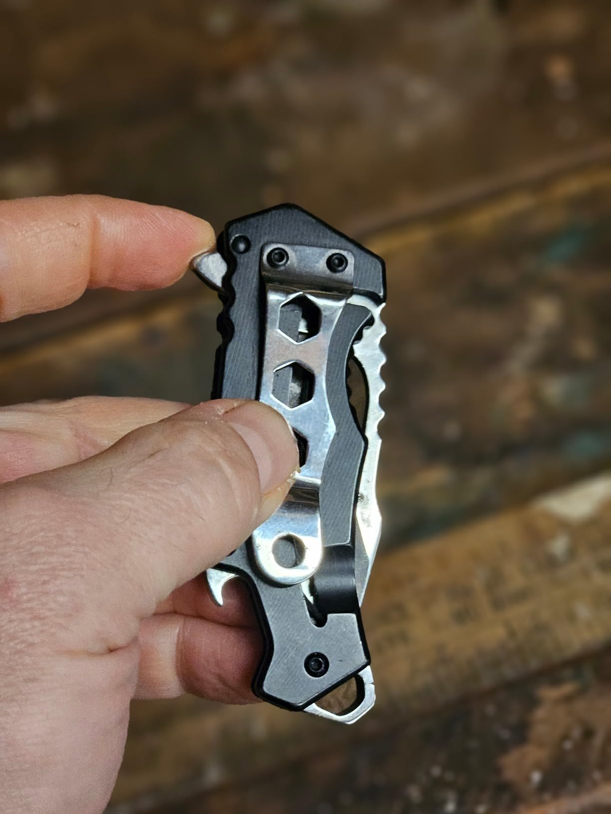Black MTech USA compact folding knife held between two fingers, clip side up, showing the hex cutouts and bottle opener