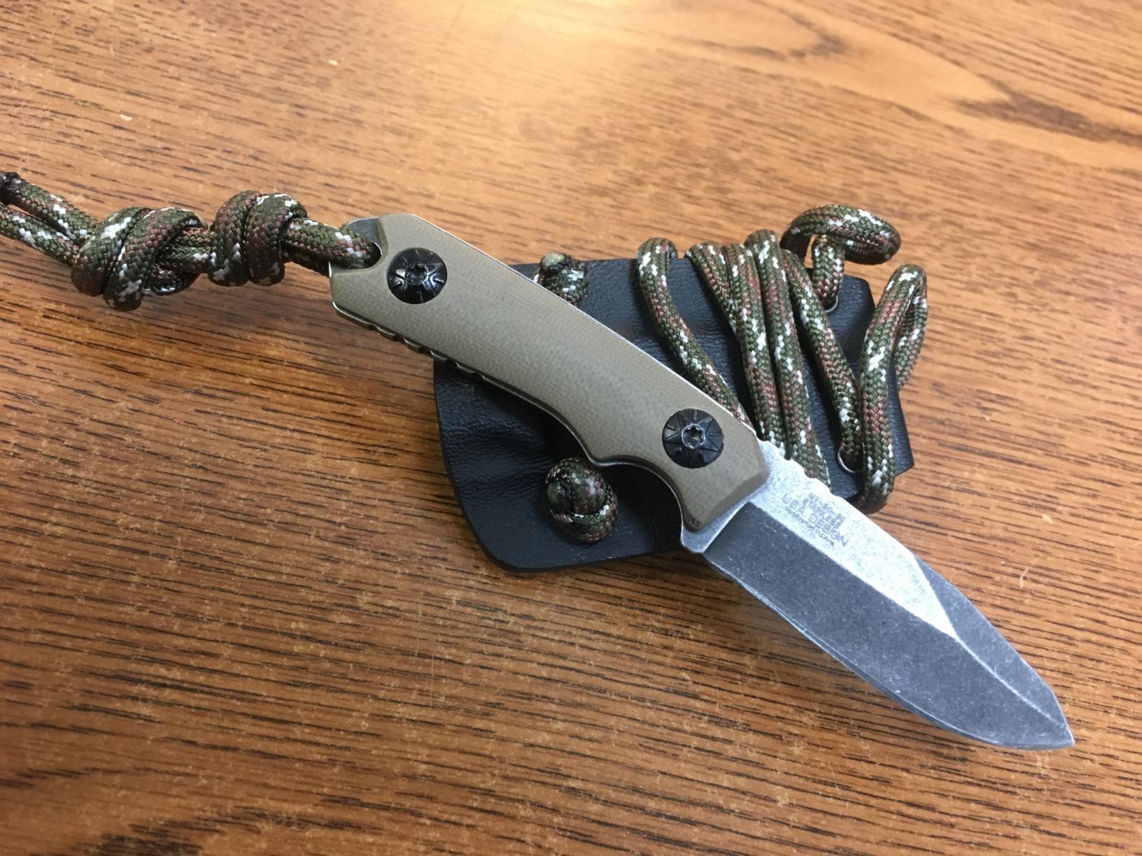 MTech USA neck knife with tan G10 scales and a stonewashed blade on an oak table, camo paracord threaded through its sheath