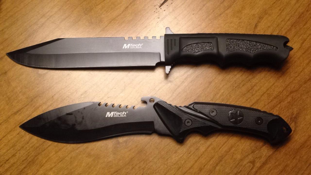Two black MTech USA fixed blades side by side on a wooden table, one with a sawback spine and one with a serrated clip point