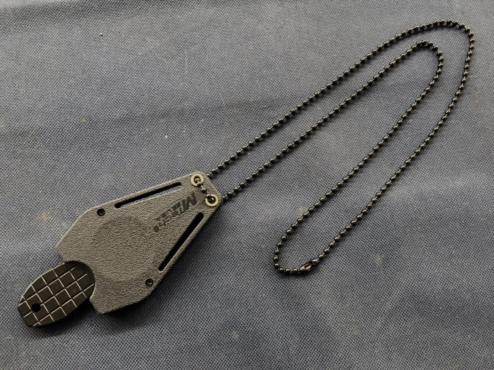MTech USA neck knife in its molded sheath on a long black ball chain, laid out on gray canvas
